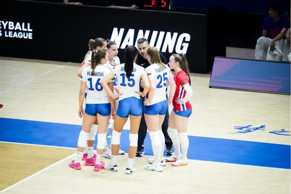 Women’s volleyball team huddles with coach on a light wood court, wearing white and blue uniforms with numbers visible (16, 15, 25).