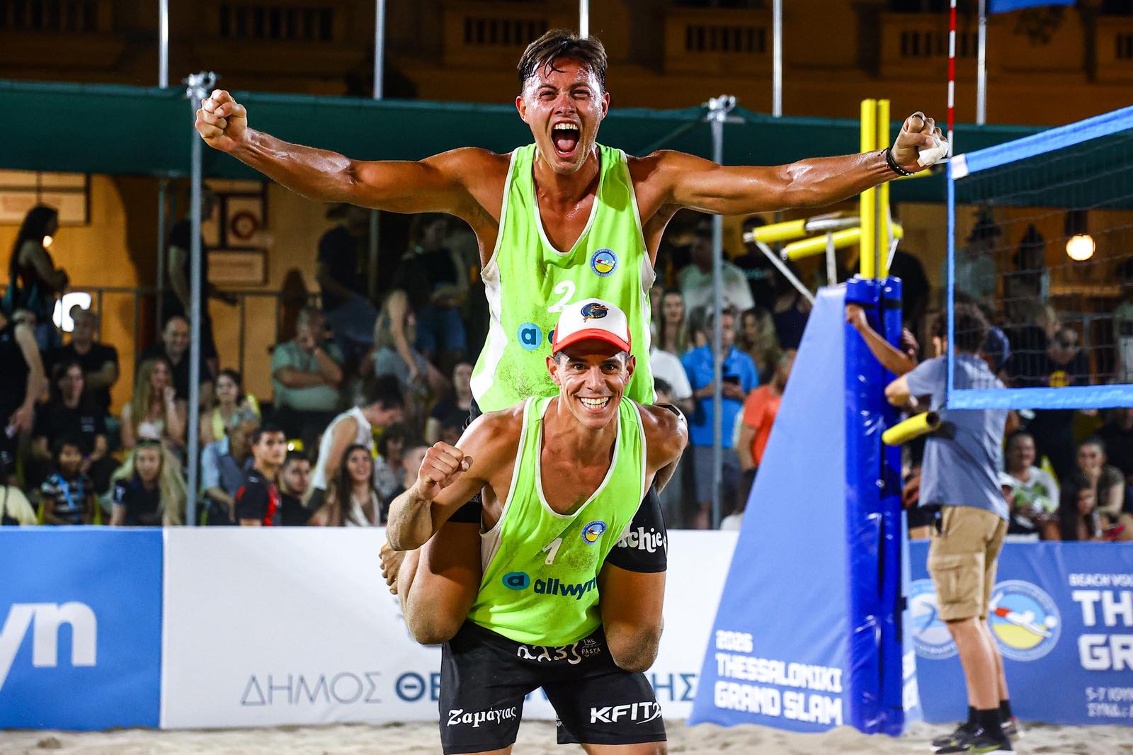 ntallaschatziwinnersthess2026 The Volley Lovers Two male beach volleyball players in neon green jerseys celebrate on the sand court as the crowd cheers in the background.