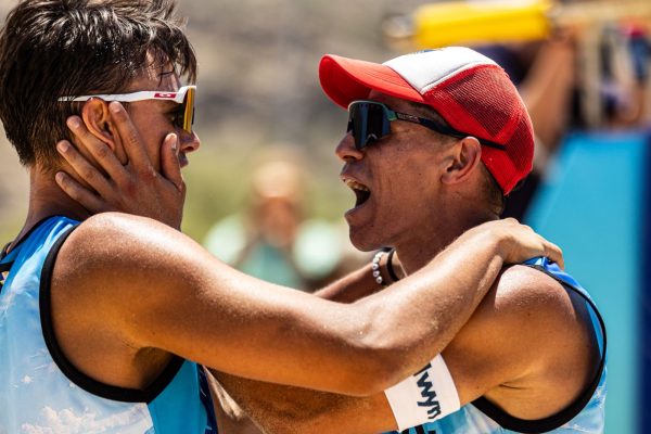 Two athletes in blue uniforms embrace, one with a red cap shouting or cheering in celebrationor encouragement, outdoor setting with blurred background.
