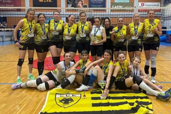 A women's volleyball team in yellow jerseys posing with medals and a trophy on a gym floor, kneeling and standing in front of a net.