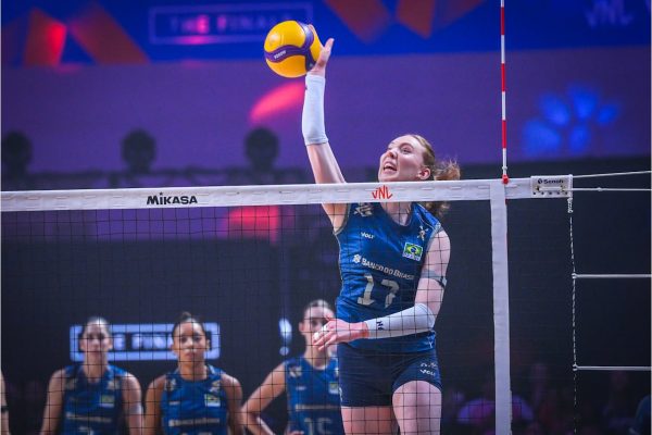 Brazilian female volleyball player in a blue uniform jumps to spike a yellow ball over the net during a match.