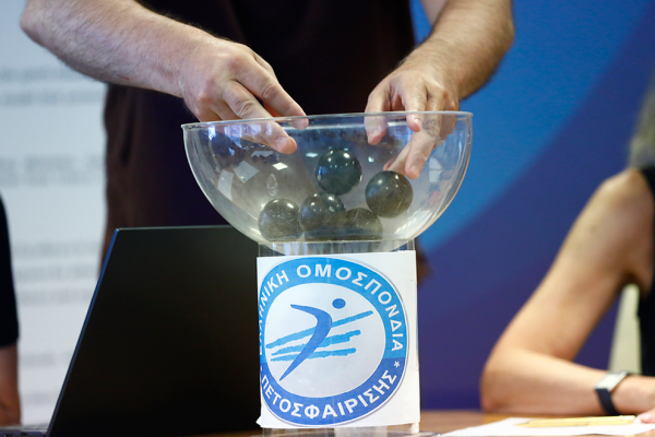 Hands reach into a glass bowl of dark balls for a raffle on a pedestal with a blue Greek federation emblem nearby.