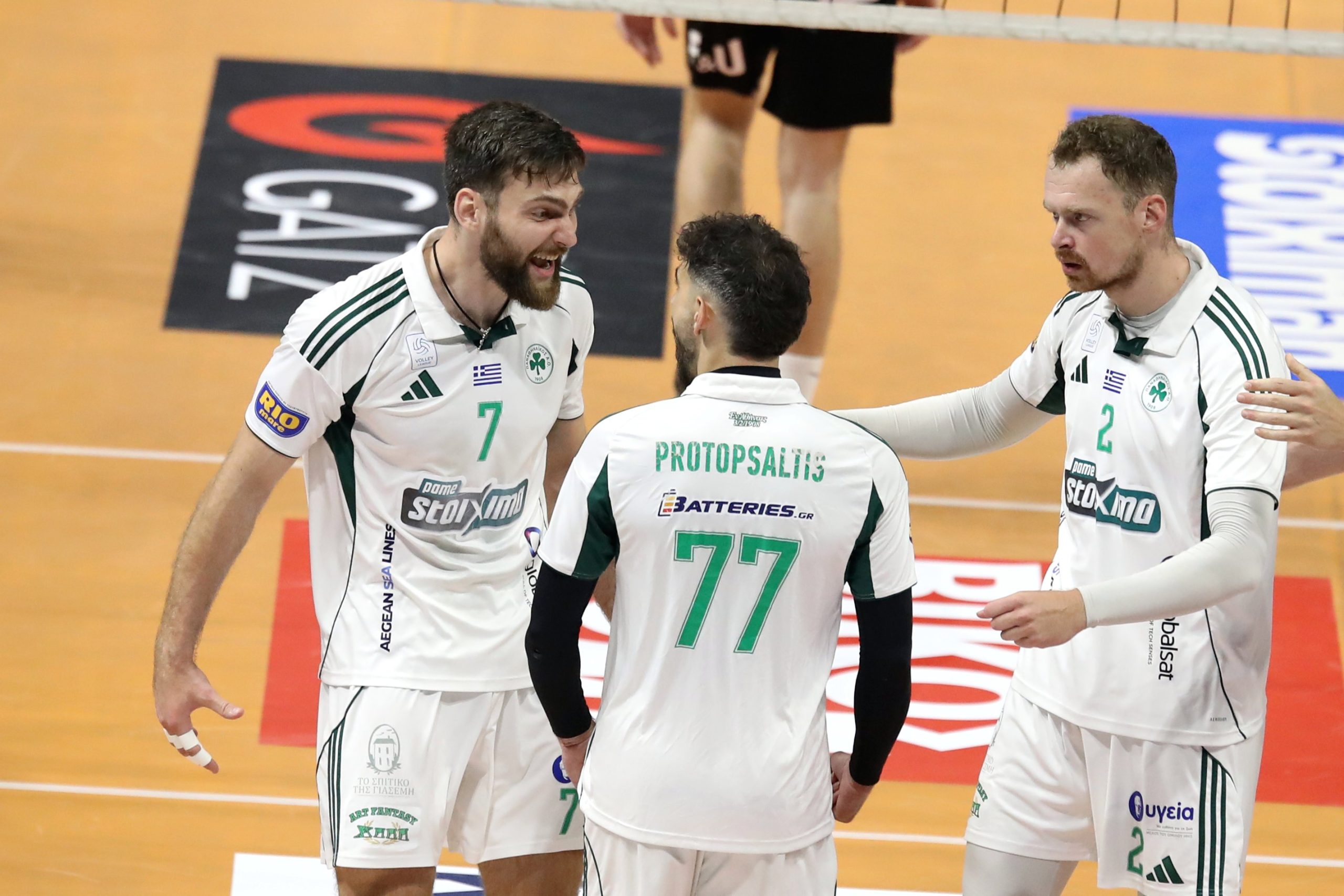Three volleyball players in white and green uniforms huddle on an indoor court, talking strategy between plays.