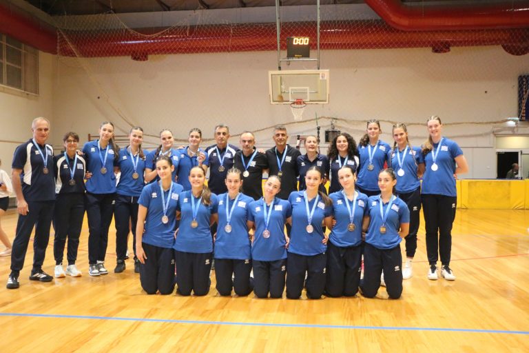 IMG7386scaled The Volley Lovers Group of athletes in blue uniforms posing in a gym, medals around their necks. In the background is a basketball hoop and gym netting.