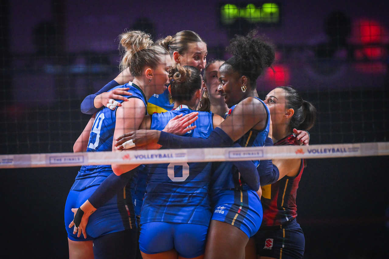 Women’s volleyball team in blue uniforms huddle together in a close group on the court, celebrating a point by the net.