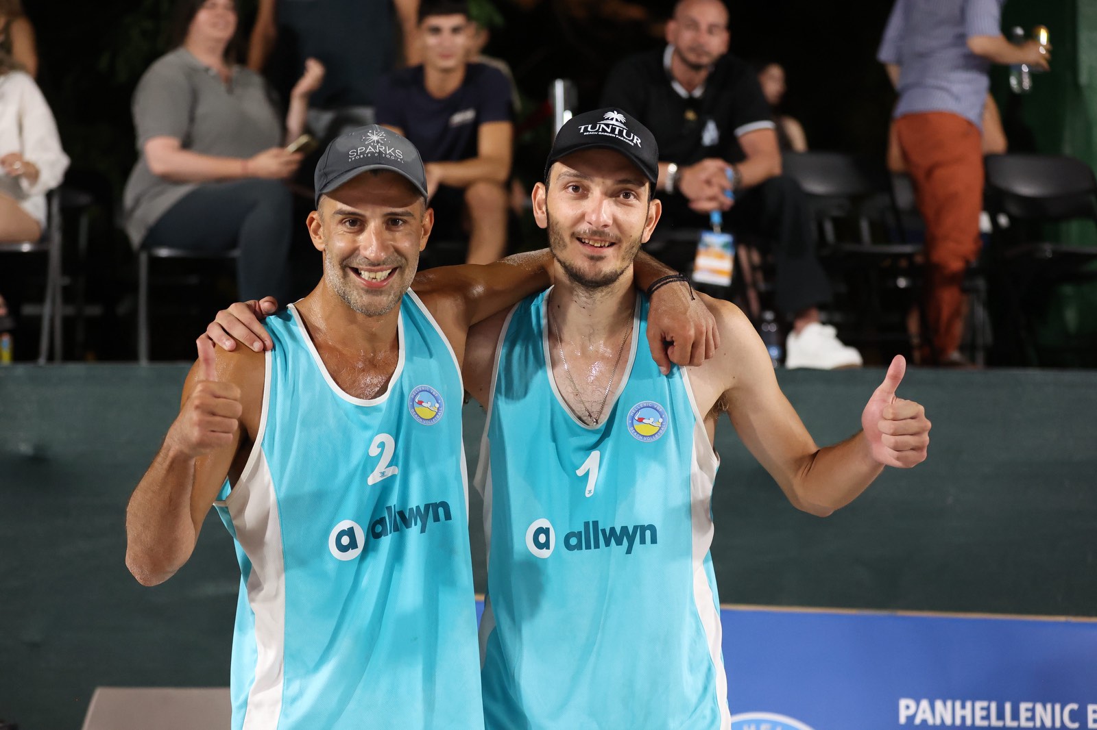 Two male volleyball players in turquoise jerseys with arms around each other, smiling and giving thumbs up after a match on the court.