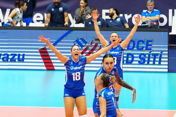 Women’s volleyball team in blue uniforms celebrates on the court with arms raised in victory.