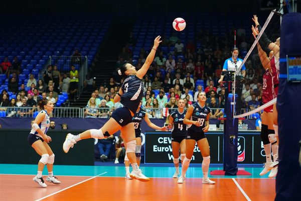 Female volleyball player in dark uniform jumps for a spike over the net as teammates and spectators look on in a crowded arena.