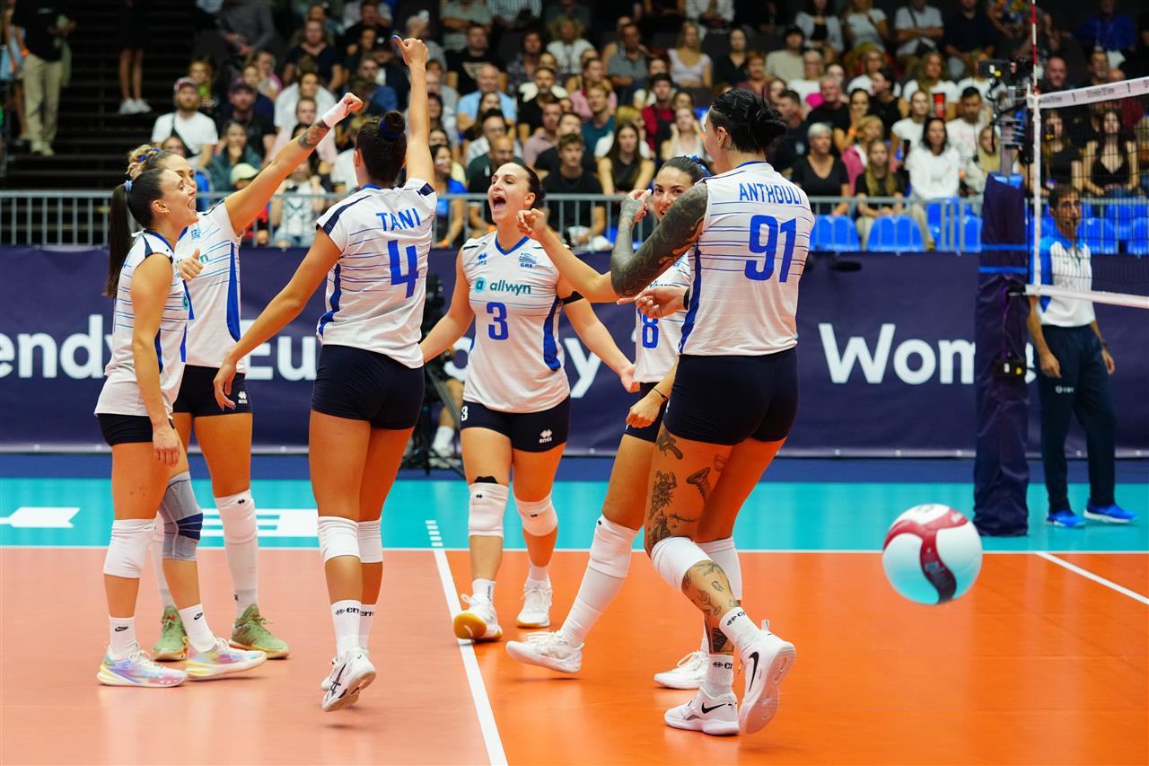 181827BUL GRE 23082026 12 The Volley Lovers Six female volleyball players in white and blue uniforms celebrate on an orange court, high-fiving as a ball rolls by.