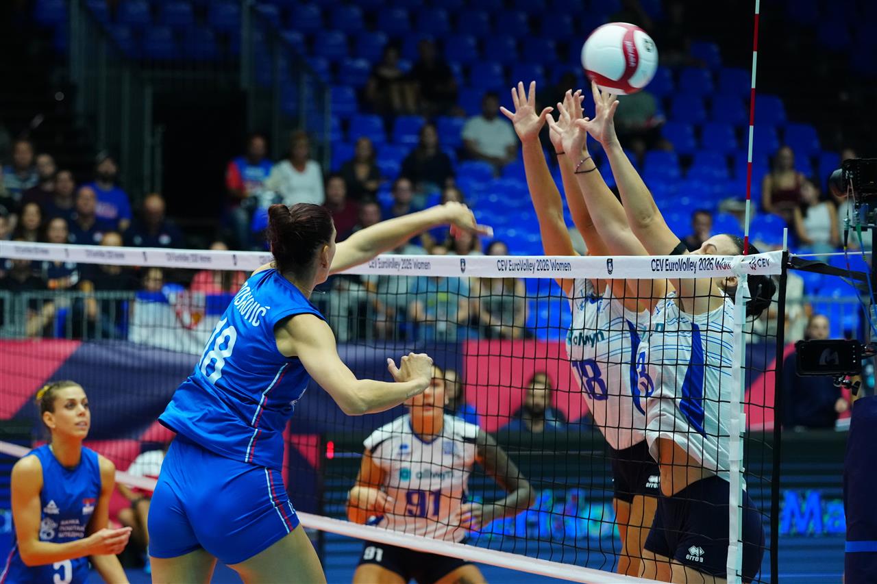 195033SRB GRE 26082026 5 The Volley Lovers Female volleyball players at the net: a blue-team attacker hits the ball while two white-team blockers jump to block.