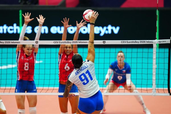 Spiker in white jersey (#91) jumps to hit the ball over the net as two red-jersey blockers leap to block.