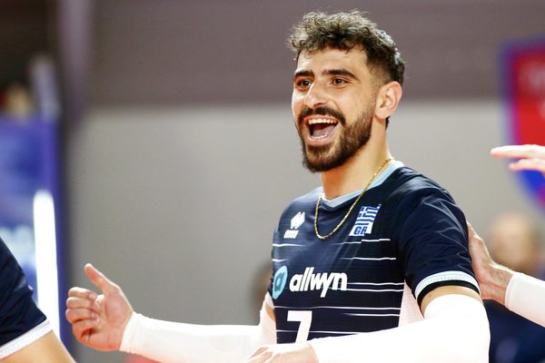 Male volleyball player celebrating a point, arms outstretched, wearing a dark blue jersey with Greek insignia and white stripes.