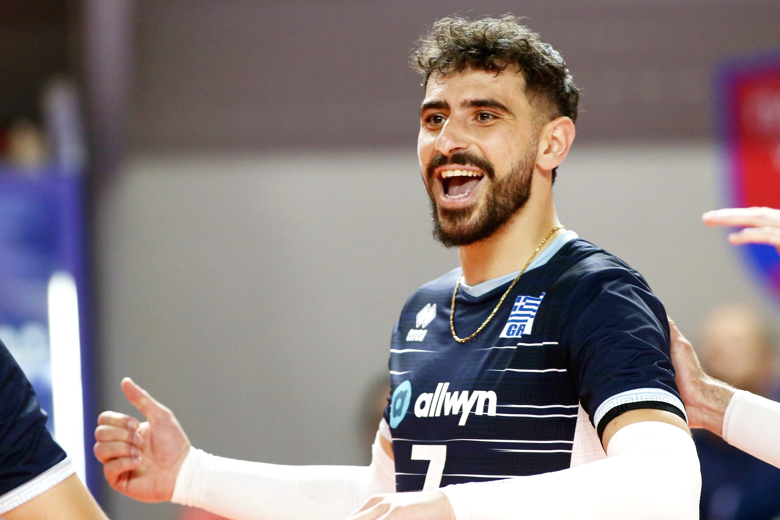 <br>ÅËËÁÄÁ ÅËÂÅÔÉÁ<br>ÖÉËÉÊÏ The Volley Lovers Male volleyball player celebrating a point, arms outstretched, wearing a dark blue jersey with Greek insignia and white stripes.
