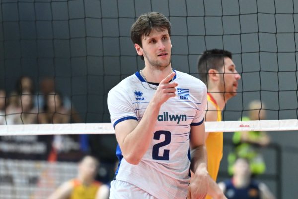 Male volleyball player in a white jersey with blue trim points toward the action, net in foreground.