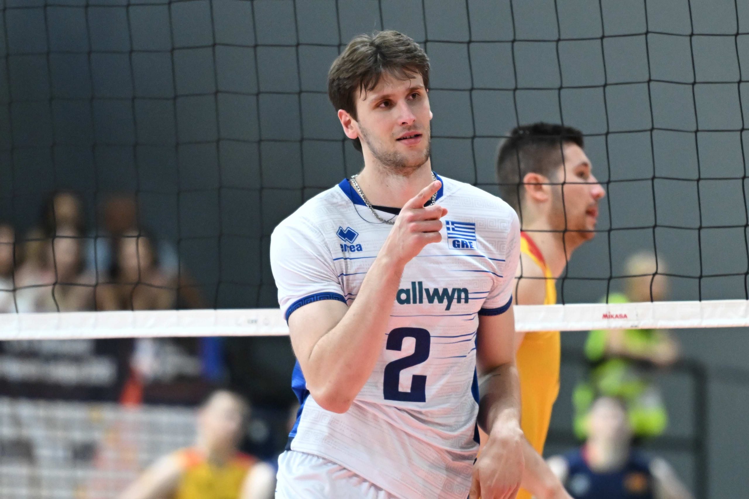 Male volleyball player in a white jersey with blue trim points toward the action, net in foreground.