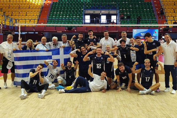 Volleyball team posing with a Greek flag in an indoor arena after a match, with Spain and Greece flags overhead and a 'Match Result' banner in the background.
