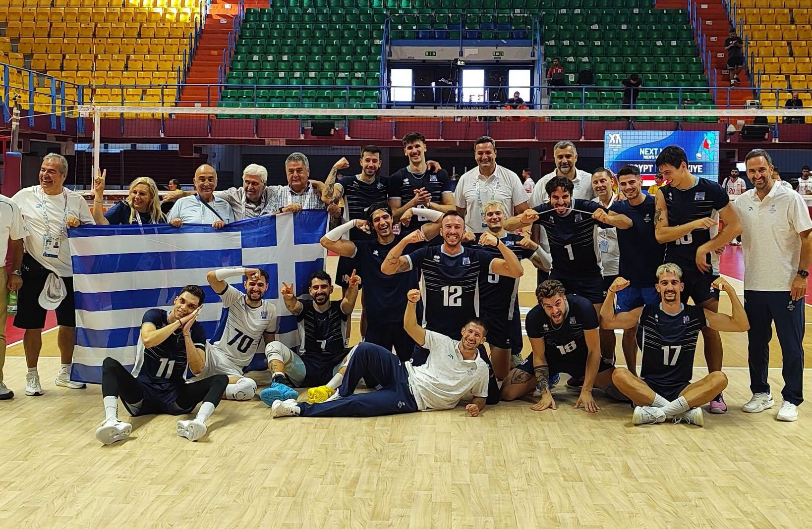 78628912810737733683804978757408268632007545n The Volley Lovers Volleyball team posing with a Greek flag in an indoor arena after a match, with Spain and Greece flags overhead and a 'Match Result' banner in the background.