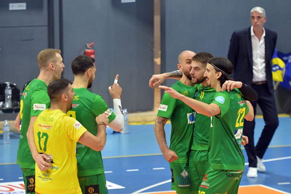Group of male volleyball players in green uniforms celebrate and high-five on an indoor blue court, with a yellow-jersey player nearby.