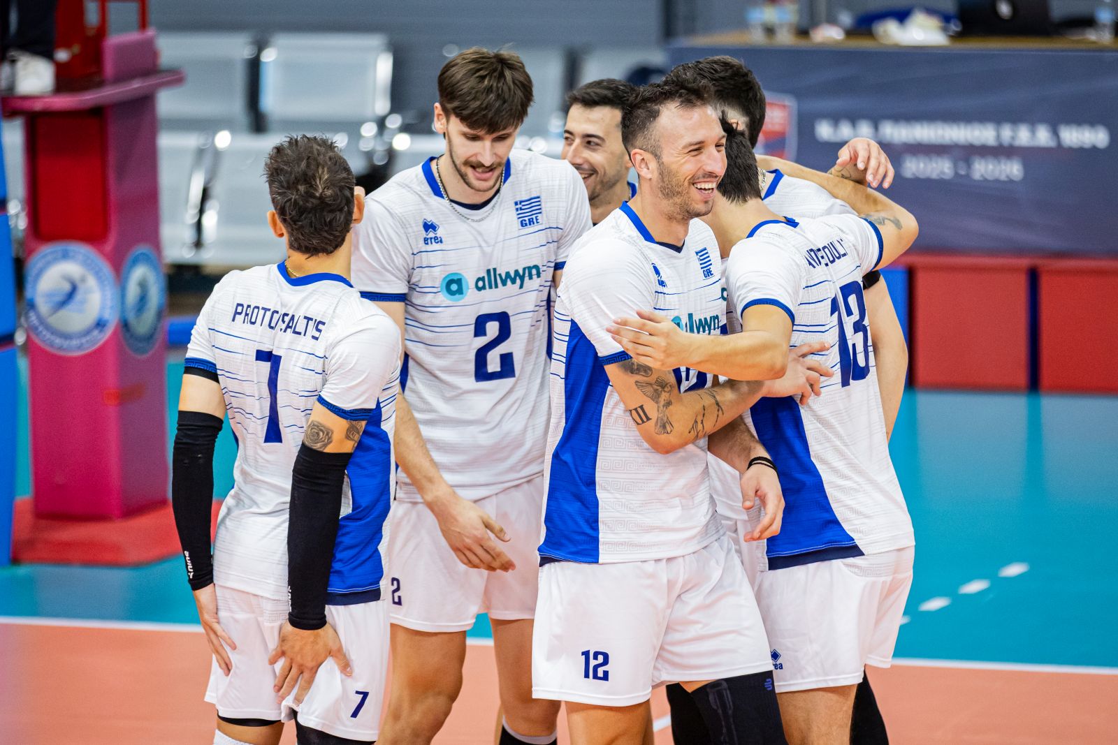 Group of male volleyball players in white and blue uniforms embrace in celebration on an indoor court.