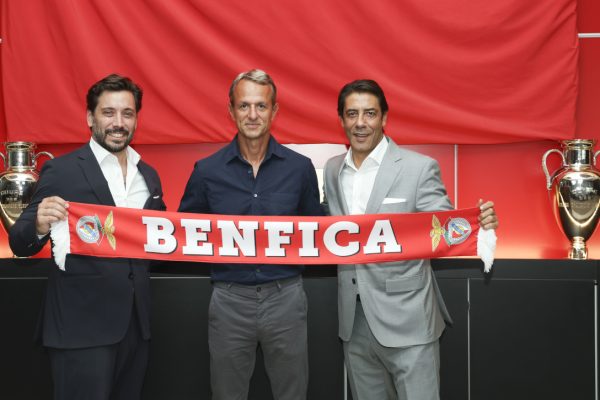 Three men in suits pose together, holding a red Benfica scarf in front of two silver trophies in a bright room.
