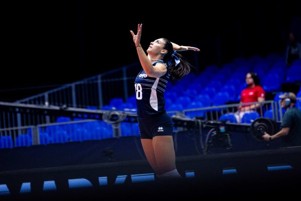 Female volleyball player in a blue uniform reaching upward with arms raised in a dark arena.