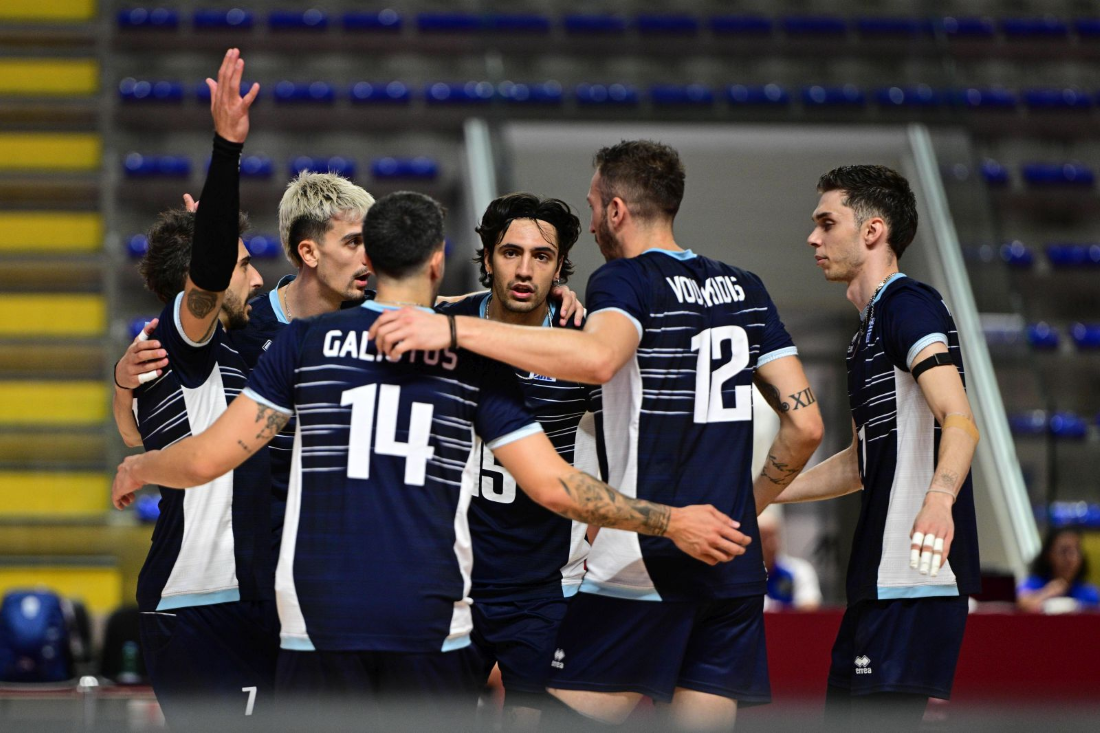 Team of male volleyball players in navy uniforms huddles together on an indoor court, number 14 and 12 visible on jerseys.