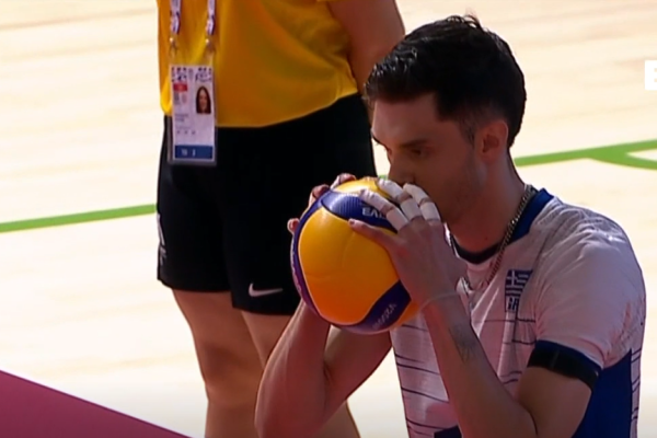 Male volleyball player in a white jersey holds a yellow and purple volleyball to his lips on an indoor court.