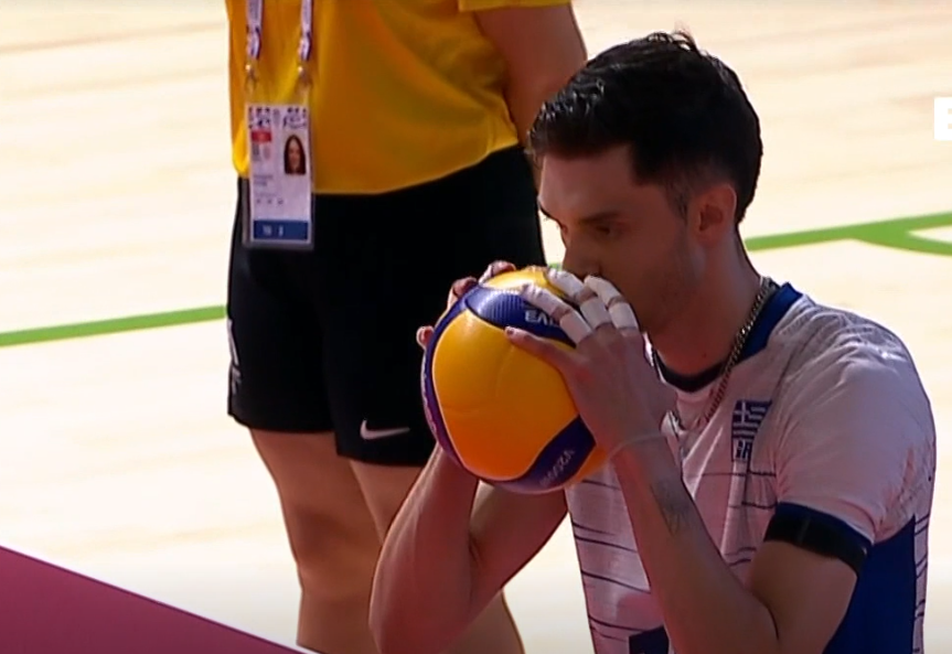 image The Volley Lovers Male volleyball player in a white jersey holds a yellow and purple volleyball to his lips on an indoor court.
