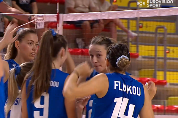 Female volleyball players in blue jerseys huddle on the court near the net during a match; jersey 14 shows the name on the back.