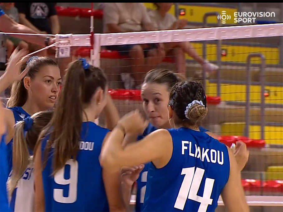 image2 The Volley Lovers Female volleyball players in blue jerseys huddle on the court near the net during a match; jersey 14 shows the name on the back.