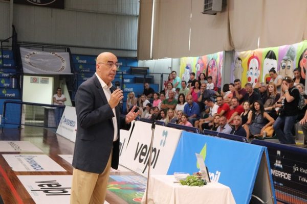 Bald man in a blazer speaks into a microphone from a table podium as a crowd watches in an indoor gymnasium with colorful banners.