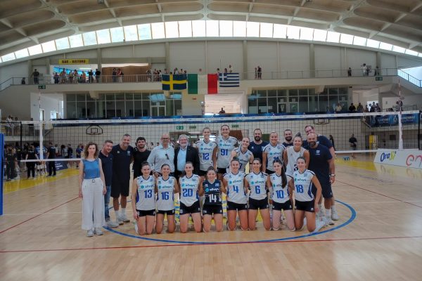 Group of volleyball players in blue-and-white uniforms posing for a team photo on an indoor court, with spectators on a balcony above and flags hanging overhead.