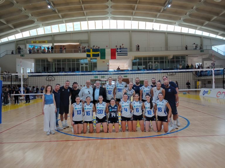 Group of volleyball players in blue-and-white uniforms posing for a team photo on an indoor court, with spectators on a balcony above and flags hanging overhead.