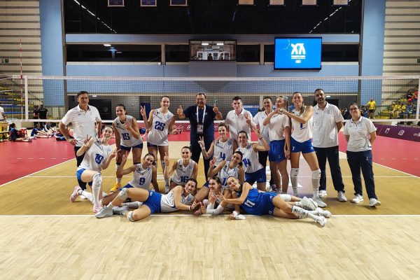 Team of female volleyball players and coaches posing for a group photo on an indoor court, cheering with victory signs near the net.