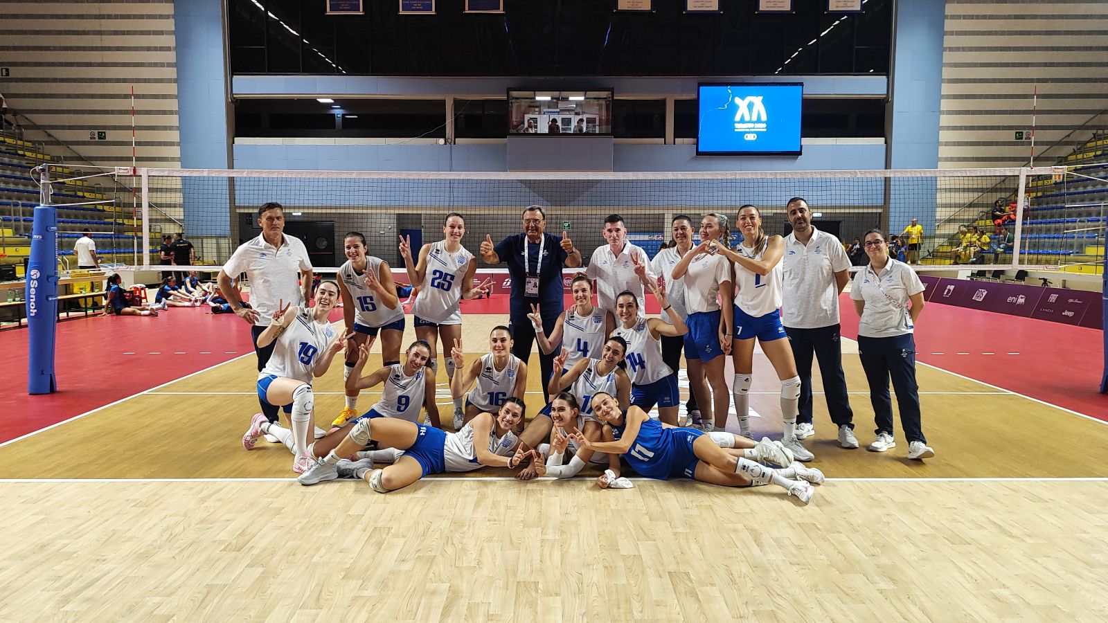 wnnt2026teamkouvelos The Volley Lovers Team of female volleyball players and coaches posing for a group photo on an indoor court, cheering with victory signs near the net.