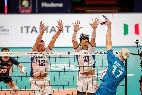 Volleyball action at the net: two players in white and blue jerseys jump to block a spike from an opponent in blue (number 17) as the ball rises above the net.