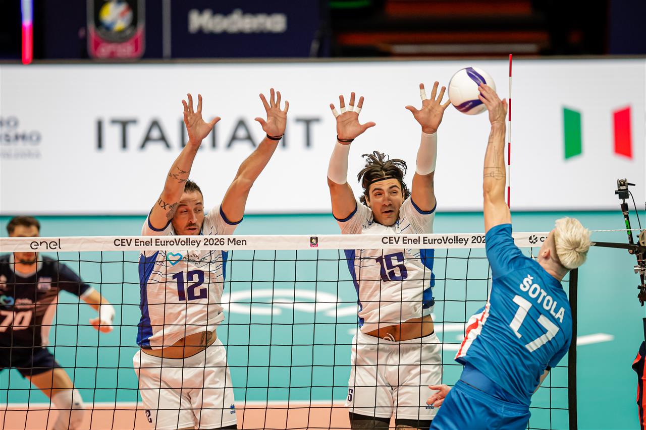 164712I8A1573 The Volley Lovers Volleyball action at the net: two players in white and blue jerseys jump to block a spike from an opponent in blue (number 17) as the ball rises above the net.