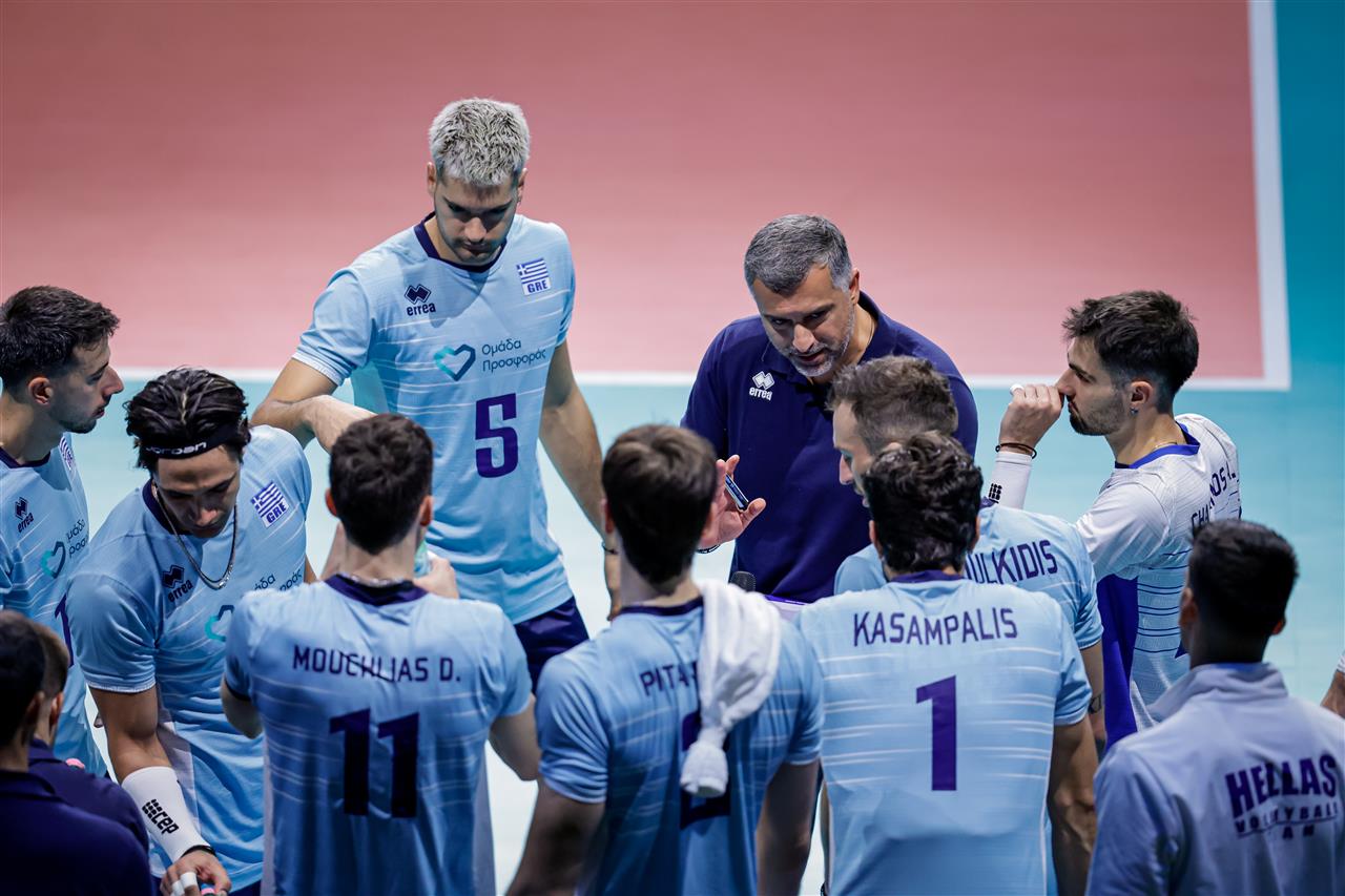 170514I8A4648 The Volley Lovers Greek men's volleyball team in light blue uniforms huddles around their coach on a blue court.