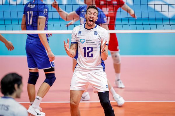 Male volleyball player in white jersey #12 shouting with hands raised at the net during a rally, blue and red opponents in background on a pink court.