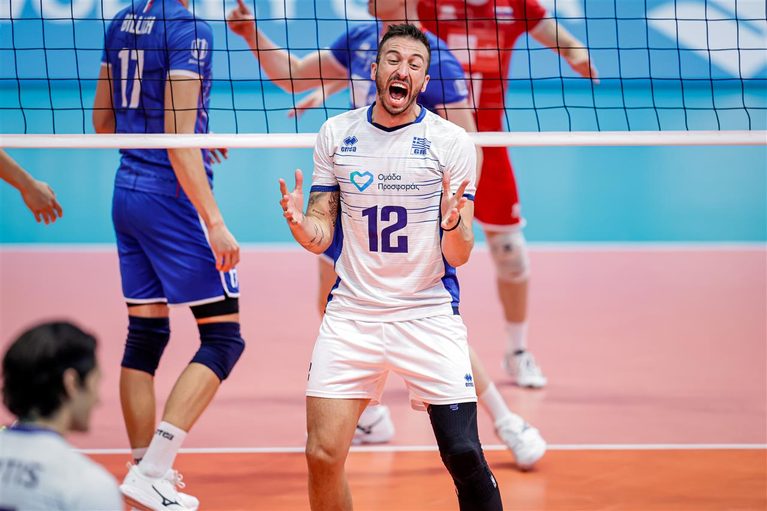 175458I8A6376 The Volley Lovers Male volleyball player in white jersey #12 shouting with hands raised at the net during a rally, blue and red opponents in background on a pink court.