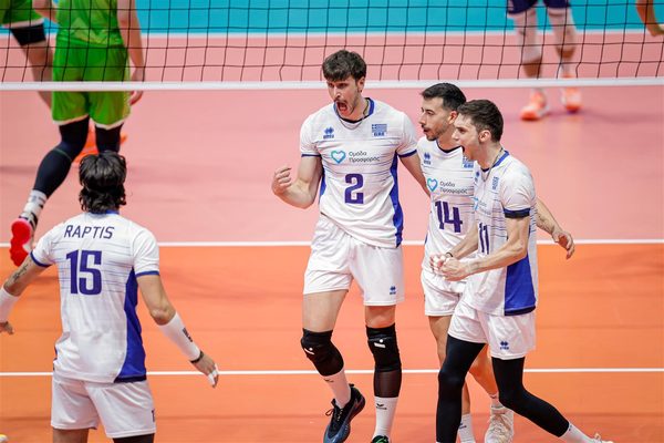 Four volleyball players in white and blue celebrate on an orange court near the net.