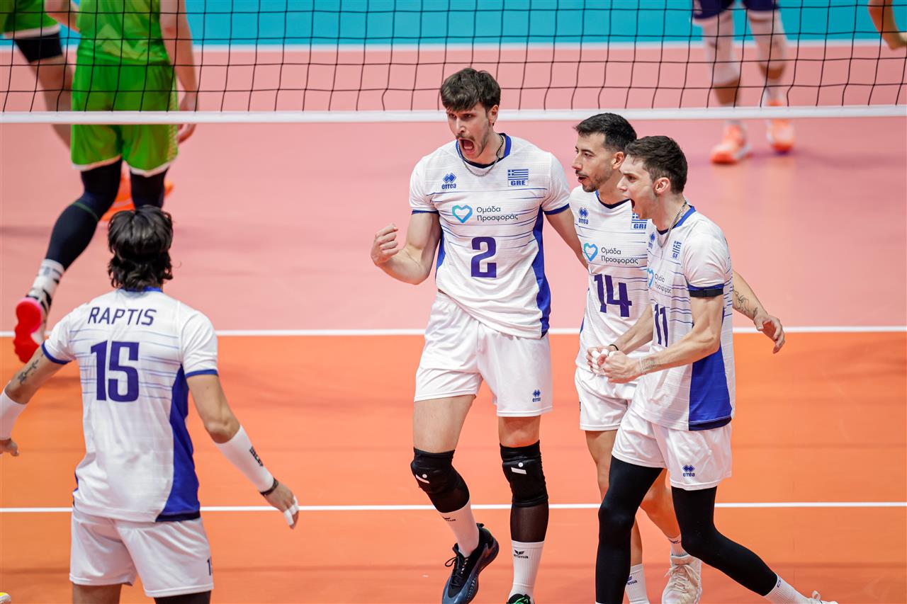 Four volleyball players in white and blue celebrate on an orange court near the net.