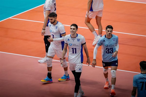Volleyball players in white and blue uniforms walk off the court, smiling after a game on an orange indoor court.