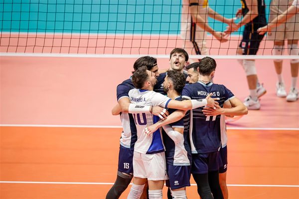 A group of male volleyball players in navy and white uniforms hug in celebration on an orange indoor court near the net.