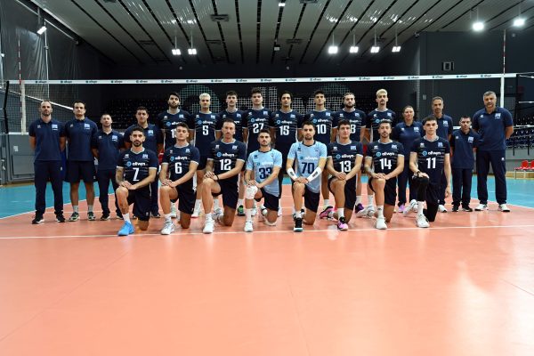 Group of volleyball players and coaches posing for a team photo in an indoor gym.