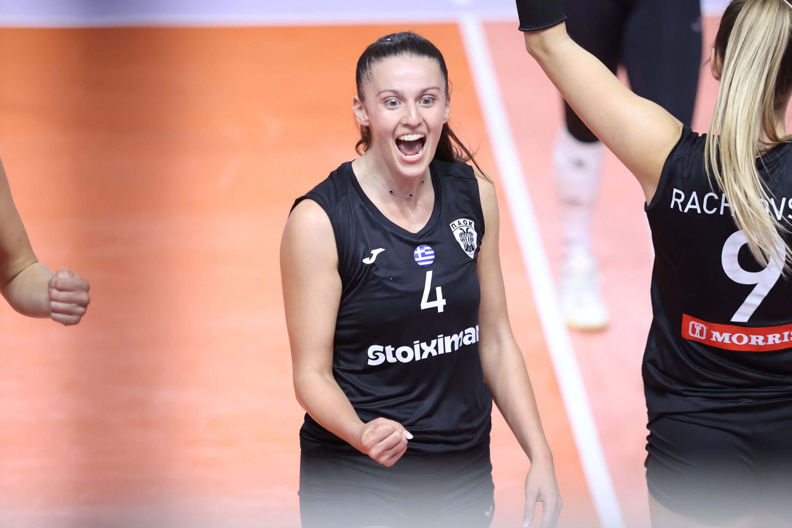 5356660 The Volley Lovers Two volleyball players in black uniforms celebrate with clenched fists on an orange court, one with number 4 on her jersey and a Greek flag pin on her chest.