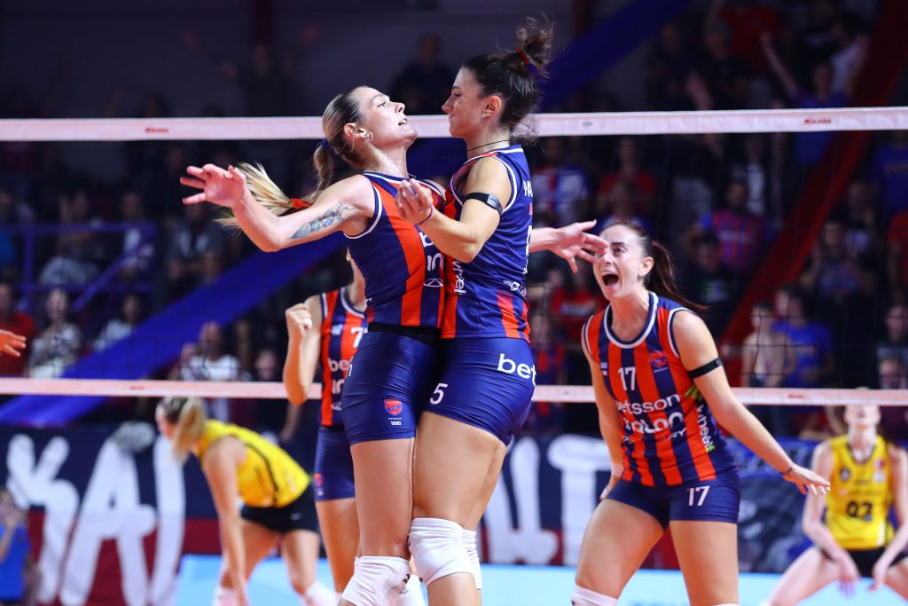 5357224 The Volley Lovers Two female volleyball players in blue‑red uniforms celebrate a point at the net, arms raised as teammates cheer in the background.