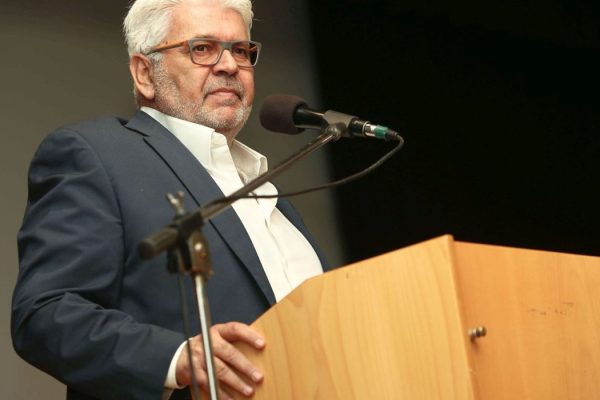 Older man with gray hair and glasses speaks at a wooden lectern into a microphone.