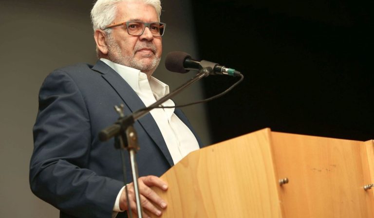 6370434621166146831200x700 The Volley Lovers Older man with gray hair and glasses speaks at a wooden lectern into a microphone.