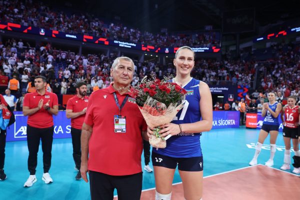 Man in red polo with badge stands beside smiling athlete in blue holding a bouquet of red roses, in a crowded indoor arena after a match.